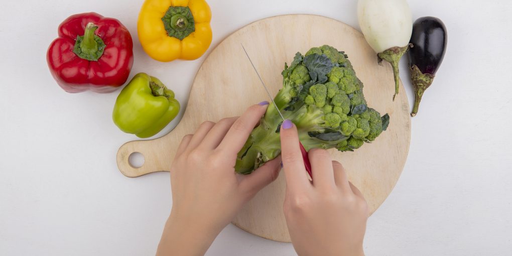 top view  woman cuts broccoli on cutting board with colored bell peppers and eggplants on white background