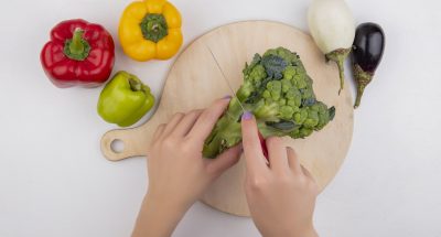 top view  woman cuts broccoli on cutting board with colored bell peppers and eggplants on white background