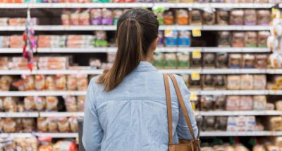 In this rear view, an unrecognizable woman stands with a shopping cart in front of a shelf full of food in the bread aisle of a grocery store.