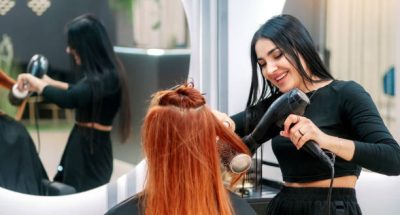 Hair styling in beauty salon. Woman does her hair in modern beauty salon. Woman stylist dries hair with hairdryer to salon client and smiles.