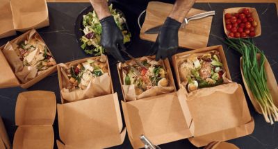Vegetables added to the burgers. Man is packing food into the paper eco boxes. Indoors, restaurant.