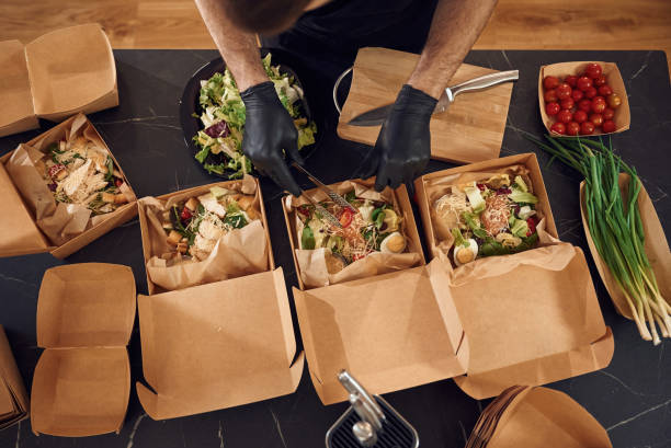 Vegetables added to the burgers. Man is packing food into the paper eco boxes. Indoors, restaurant.