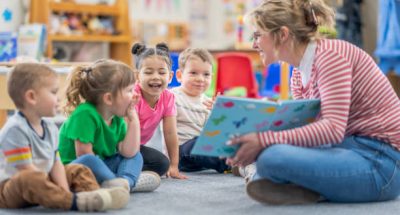 A preschool teacher sits on the floor of her classroom with a small group of students as she reads them a book.  The children are each dressed casually and are focused on the story.