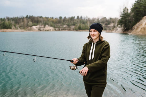 Woman catching a fish, pulling rod while fishing. Girl fishing from pier on lake or pond with text space. Fisherman with rod, spinning reel on river bank. Fishing for pike, perch, carp. Wild nature.