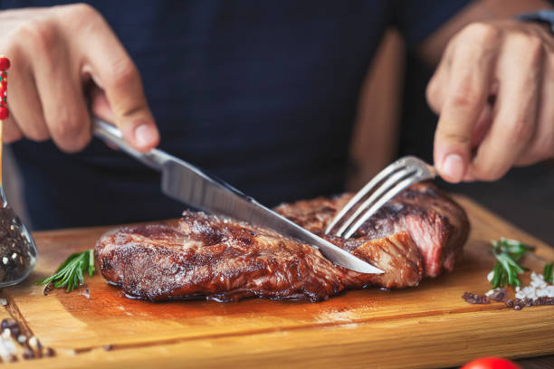 Hands holding fork and knife and eating delicious juicy steak on a wooden plate